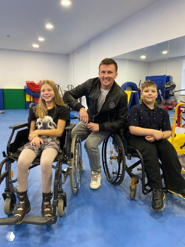 Мужчина позирует между двумя детьми в спортивном зале: оба на специальных колясках, адаптированных для танцевально-акробатических тренировок и выступлений.