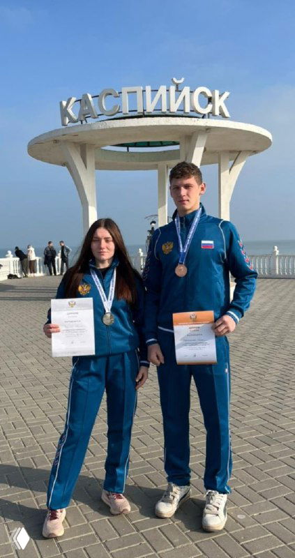 Двое рязанских кикбоксеров в спортивной форме с медалями и грамотами на фоне арки с надписью «КАСПИЙСК» у каспийской набережной.