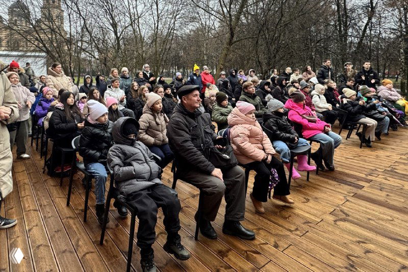 Зрители на деревянной площадке перед новой уличной сценой: взрослые и дети в тёплой одежде сидят на стульях и следят за программой на открытом воздухе.