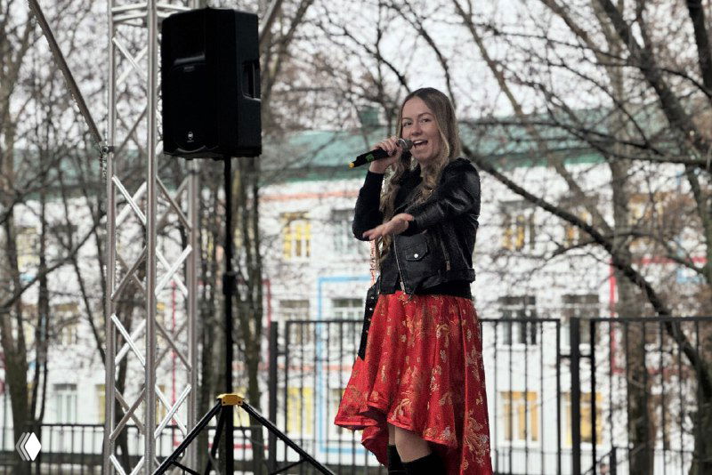 Солистка в красной юбке выступает на новой уличной сцене Дворца молодёжи, видны микрофон, колонка и городской фон за ограждением площадки.