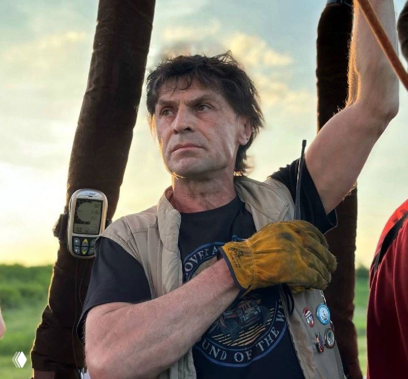 Портрет Евгения Рыбакова на открытом воздухе: мужчина в жилете и рабочих перчатках держит снаряжение и смотрит в сторону.