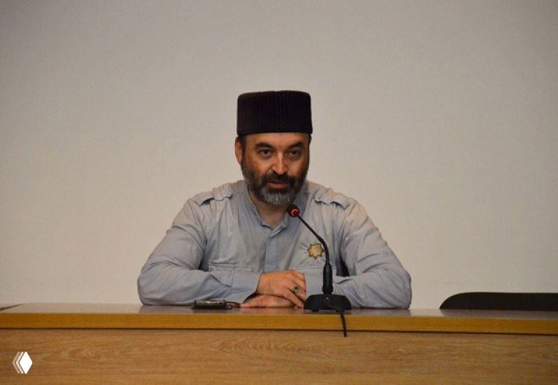 Человек в феске и светлой рубашке говорит в микрофон за столом на официальном мероприятии в Рязанской области