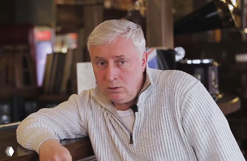 Портрет взрослого мужчины с седыми волосами в светлом свитере в интерьере заведения; видны стол, мебель и барная стойка, иллюстрация к материалу.