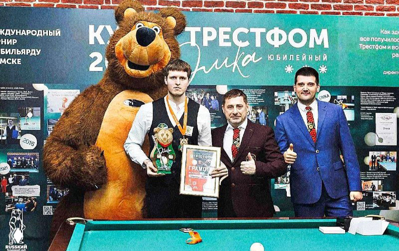 Кадр с награждения: Кирилл Гранкин в центре с серебряной медалью и кубком у бильярдного стола, рядом тренер и талисман.