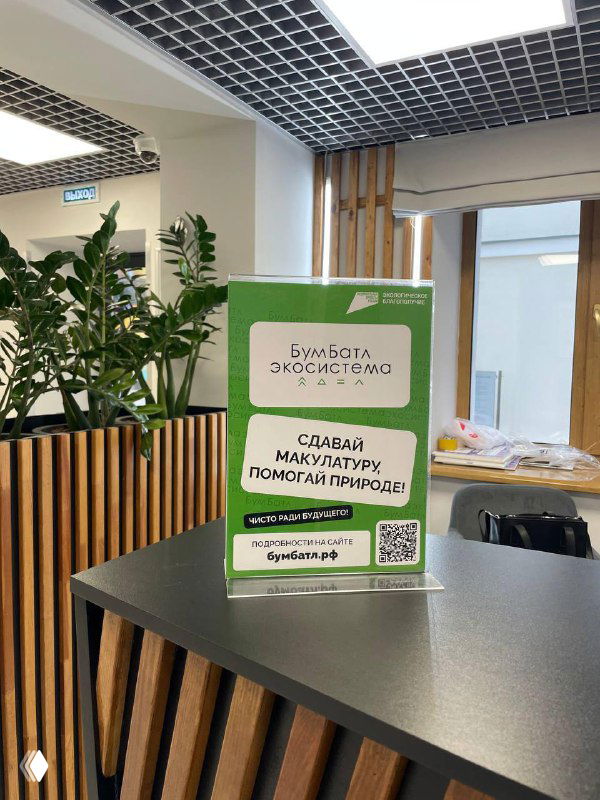 Фото информационного плаката пункта приёма макулатуры в Доме молодёжи Рязани на столе: зелёная табличка, интерьер приёмной зоны и растения на заднем плане.