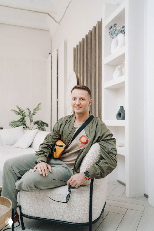 Мужчина в повседневной одежде сидит в кресле в светлом помещении с декором и растениями; нейтральный портрет для деловой заметки о продажах.