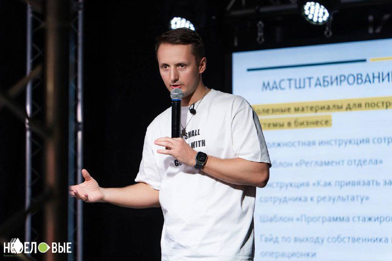 Человек на сцене с микрофоном во время деловой презентации; на заднем плане видна проекция с текстом и слайдом, деловая обстановка и аудитория