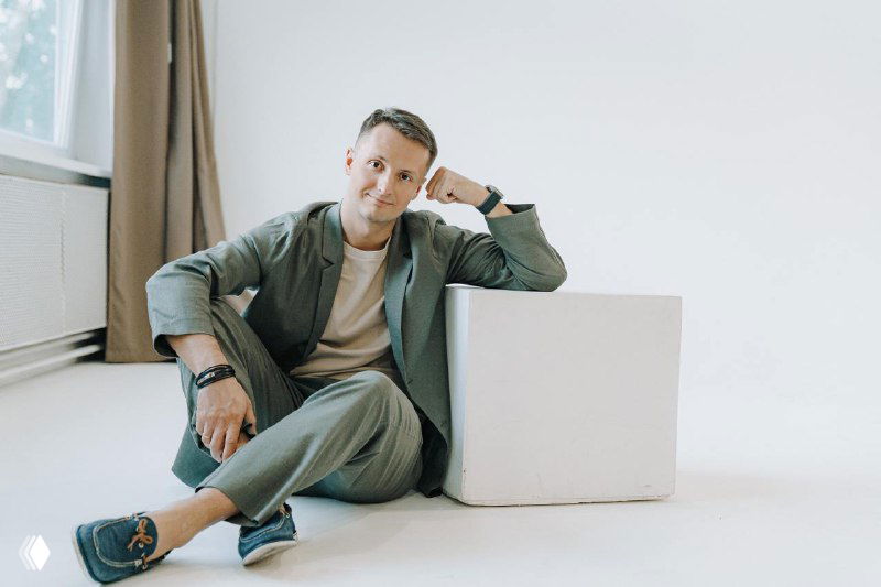 Портрет мужчины в повседневном костюме, сидящего рядом с белым кубом в светлом помещении; спокойная деловая поза и нейтральный взгляд.