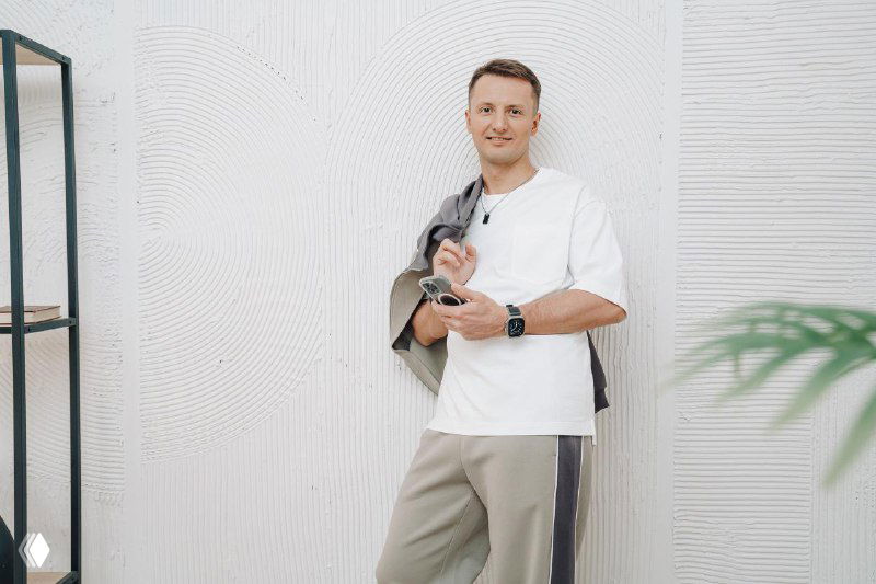 Мужчина в светлой повседневной одежде с расслабленной улыбкой у белой текстурной стены — иллюстрация к теме мотивации и продаж в команде