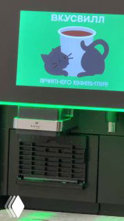 Крупный план экрана с иллюстрацией чашки и подписью «ПРИЯТНОГО КОФЕПИТИЯ», внизу части кофемашины и интерьер офисного пространства.