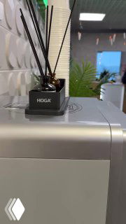 Аромадиффузор HOGA установлен на корпусе кофемашины в офисе: видна маленькая ёмкость с палочками и часть рабочего пространства на заднем плане.