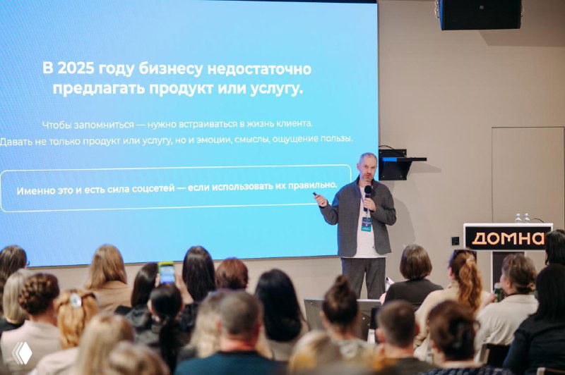 Спикер на сцене перед наполненным залом слушателей, на экране слайд про бизнес и контент; на пюпитре видна табличка «ДОМНА»