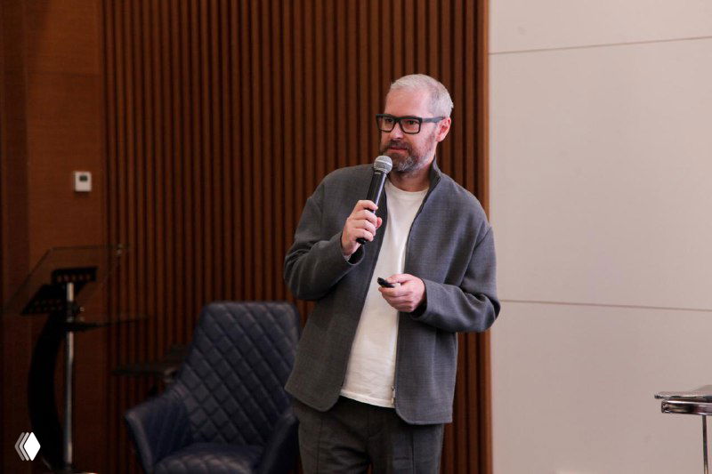 Автор канала выступает с микрофоном на сцене во время доклада на конференции; в кадре видны кресла, деревянная отделка стены и часть зала.