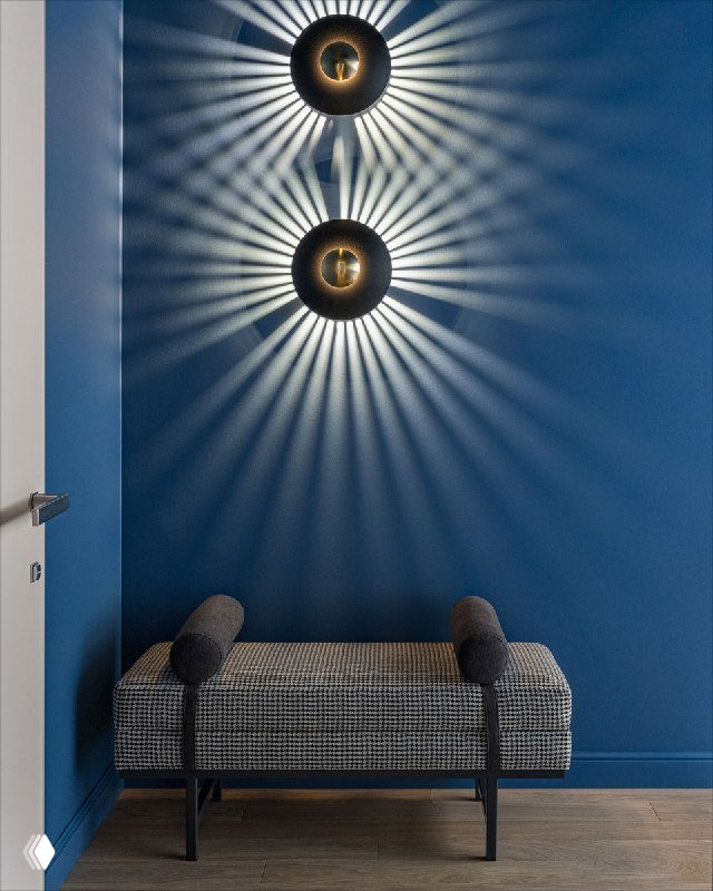 Банкетка в текстильной обивке на фоне глубокого синего коридора; над ней два круглых настенных светильника‑«солнца», отбрасывающие лучистые тени, создающие декоративный эффект.