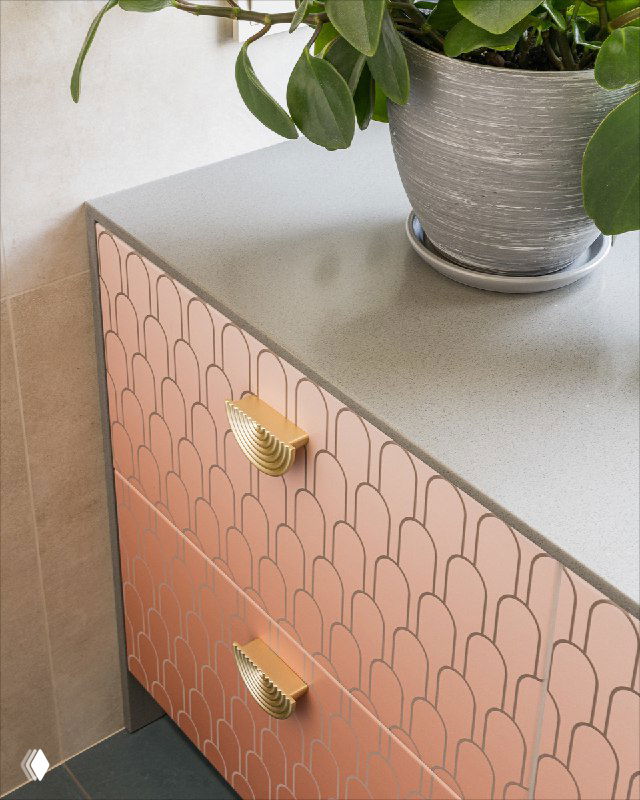 Деталь тумбы в ванной с градиентной цветной печатью фасадов и золотистыми ручками; на столешнице виден текстурный искусственный камень и декоративный горшок.