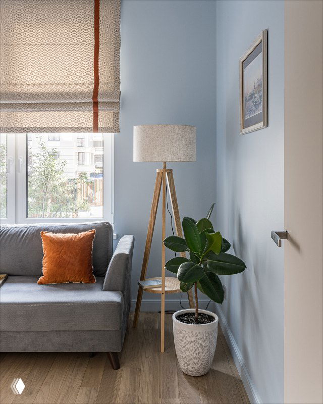 Угол гостевой комнаты: раскладной серый диван с оранжевой подушкой, акцентная римская штора, торшер с льняным абажуром, фикус и встроенный шкаф для техники.