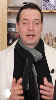 Портрет мужчины в студии интерьера: он говорит на камеру, в шарфе с микрофоном, на фоне полки с образцами и декором.