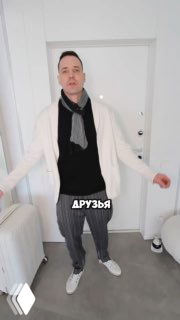 Мужчина в светлом кардигане и шарфе стоит внутри квартиры у входной двери; внизу кадра заметна наклейка с текстом «ДРУЗЬЯ», интерьер в нейтральных тонах.