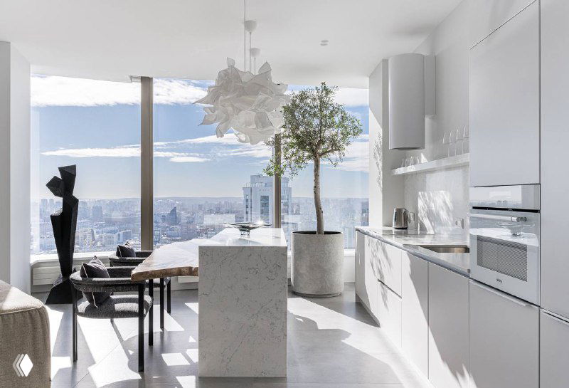 Широкий вид белой альпийской кухни в апартаментах Башни Исеть: остров-стойка, белый кварцевый агломерат, панорамные окна и городской пейзаж за ними.