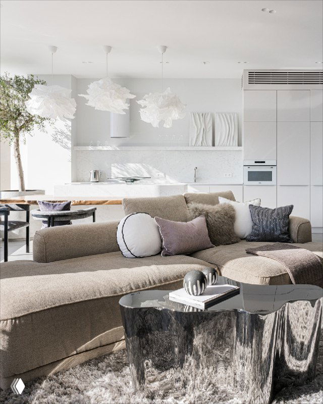 Мягкая зона с большим бежевым изогнутым диваном, дизайнерскими зеркальными столиками и открытой светлой кухней на заднем плане в проекте интерьера.