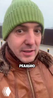 Крупный план мужчины в зелёной вязаной шапке и коричневой куртке на улице во время поездки в Казань; видеокадр с надписью «РЕАЛЬНО».