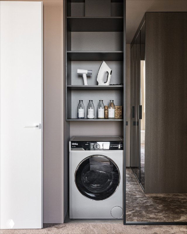 Ниша в гардеробной с фронтальной стиральной машиной, скрытой за зеркальной панелью; видны полки с моющими средствами и ковровое покрытие, плавно переходящее в спальню.