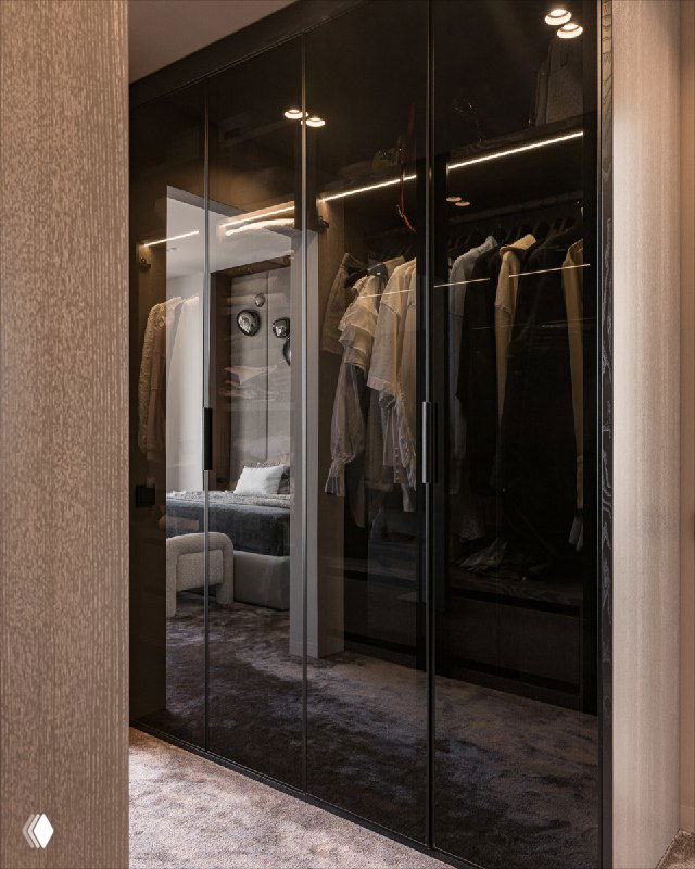 Тонированные стеклянные фасады шкафов гардеробной с внутренней подсветкой; отражение спальни в зеркале и аккуратно развешанная одежда создают элегантный интерьер.