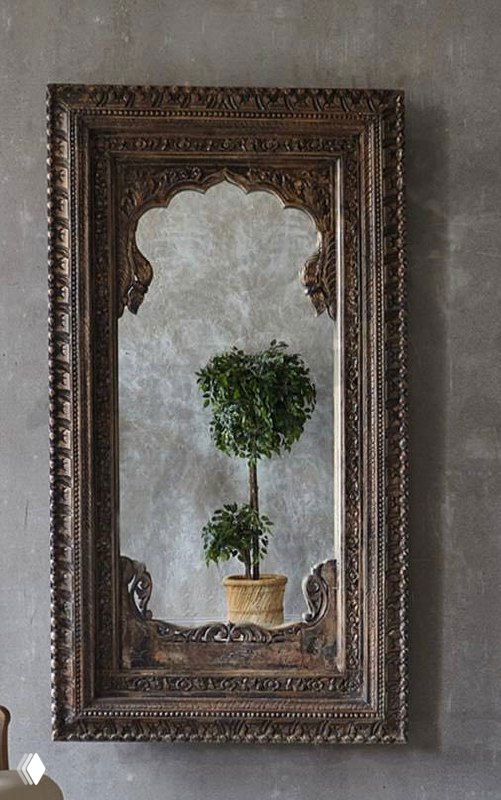 Детальное фото резного зеркала в массивной деревянной раме на фактурном фоне, перед ним небольшое декоративное растение в горшке.