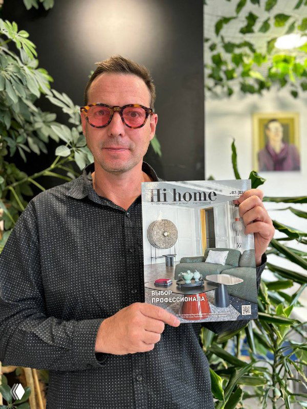 Мужчина в очках держит журнал Hi home с обложкой роскошного интерьера проекта клубного дома «Кандинский», на фоне зелёных растений.