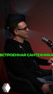 Мужчина в подкаст-студии у микрофона в тёмной комнате; на изображении видна зелёная надпись про встроенную сантехнику, атмосфера интервью и обсуждения дизайна.