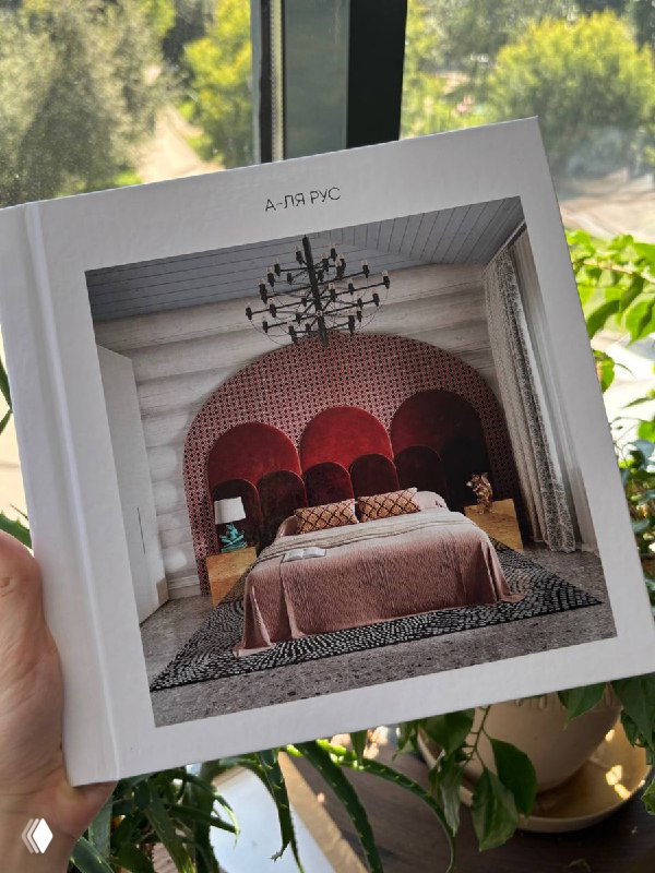 Рука держит книжку-портфолио с фотографией спальни в доме из бревна: арочное изголовье, бархатное покрывало и декоративная люстра в интерьере.
