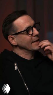 Крупный план мужчины в очках и чёрной одежде в студии; кадр из интервью или подкаста, задумчивый профиль и нейтральный фон.