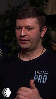 Крупный план мужчины в студии подкаста: тёмная футболка с надписью «LAZAROS PRO», жест рукой, фон с растениями и тёмной комнатой.