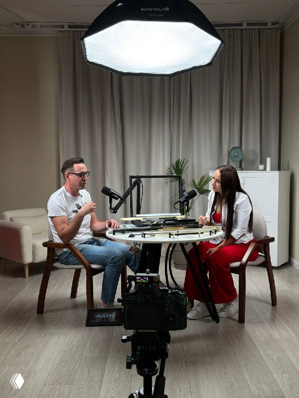 Мужчина и женщина в студии проводят подкаст за круглым столом: микрофоны, камера на штативе и софтбокс в интерьере.