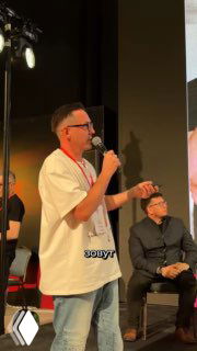 Докладчик в белой футболке с микрофоном на сцене выставки «Русский дом», рядом сидят участники, видны прожекторы.