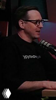 Человек в студии подкаста говорит в микрофон, в очках и тёмной одежде; видно студийный микрофон и часть интерьера записи интервью.