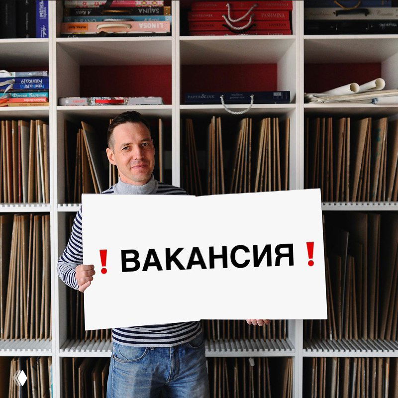 Мужчина в офисе студии дизайна держит табличку с надписью «ВАКАНСИЯ» на фоне стеллажей с образцами материалов — иллюстрация вакансии PR‑менеджера.