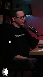 Кадр подкаста: мужчина в студии за микрофоном сидит за столом в тёмной одежде, полутёмная комната с лампой и полками на фоне, фрагмент выпуска про напольные покрытия.