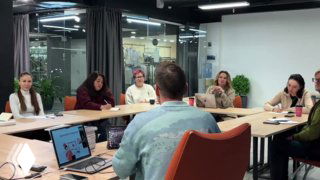 Группа слушателей и преподаватель за столом в офисной комнате на занятии по дизайну интерьера: ноутбук, блокноты и обсуждение проекта в практике курса.