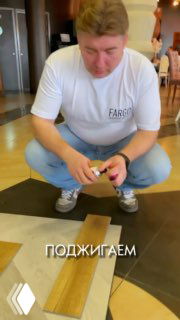 Мужчина в белой футболке с логотипом Fargo наклоняется над образцами кварц‑винилового ламината в шоуруме, демонстрирует испытание материала.