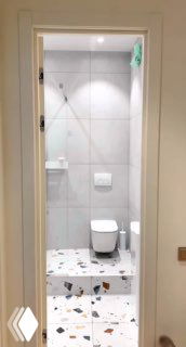 Небольшой светлый санузел с унитазом на общем подиуме и совмещённой душевой зоной; видно терраццо-покрытие пола и компактную планировку.