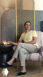 Мужчина в светлой футболке сидит в кресле в современной гостиной у декоративного очага и стеновой панели, виден интерьер зоны отдыха