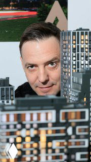 Крупный план лица дизайнера на фоне макета жилого комплекса «Архитектон», видны миниатюрные дома и фасады