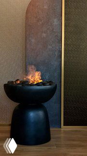 Черный декоративный напольный электрокамин в форме вазы с искусственным пламенем на фоне текстурной стены и деревянного пола.