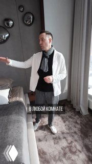 Дизайнер в светлом интерьере в белом пиджаке показывает план освещения комнаты: рядом диван, ковёр, шторы и спокойный потолок без массивной люстры.