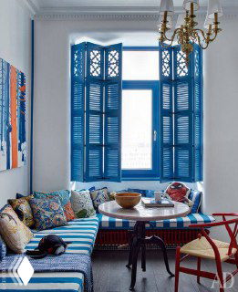 Интерьерная фотография: окно со синими деревянными ставнями, уютный обеденный уголок у подоконника с яркими подушками и декоративными картинами.