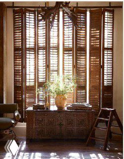 Тёплый интерьер с массивными темными деревянными ставнями, сквозь которые пробивается солнечный свет; перед окном декоративный сундук и зелень.