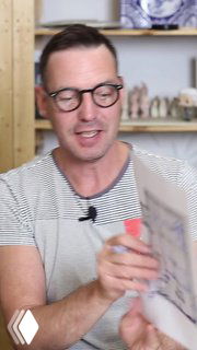 Превью к видео: автор в очках держит лист с эскизом на фоне полок и декора; кадр подчёркивает тему перепланировки и дизайн‑процесса.