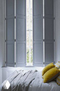 Спальная зона с высокими светло-серыми ставнями, мягкое одеяло и жёлтые декоративные подушки создают спокойный и современный образ окна.