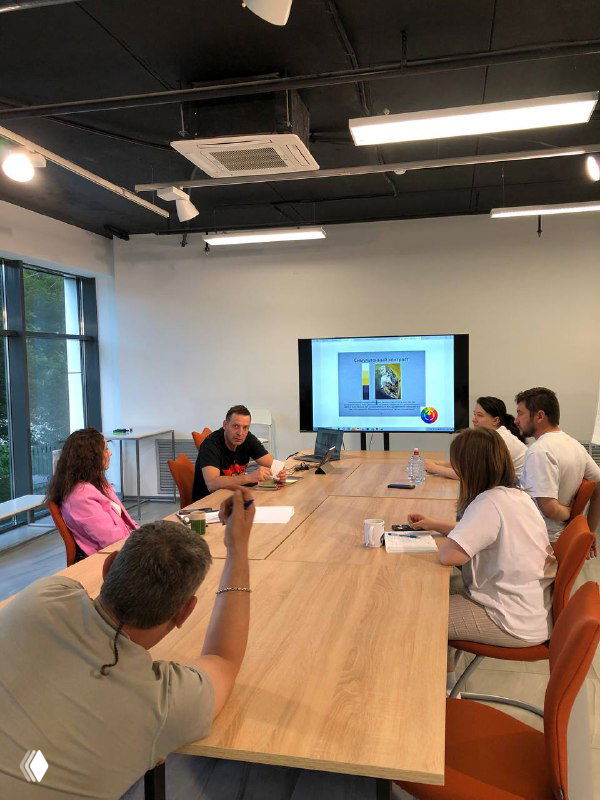 Учебная аудитория курса по дизайну интерьера: несколько человек за длинным деревянным столом, преподаватель у экрана с цветовым кругом и наглядными примерами.